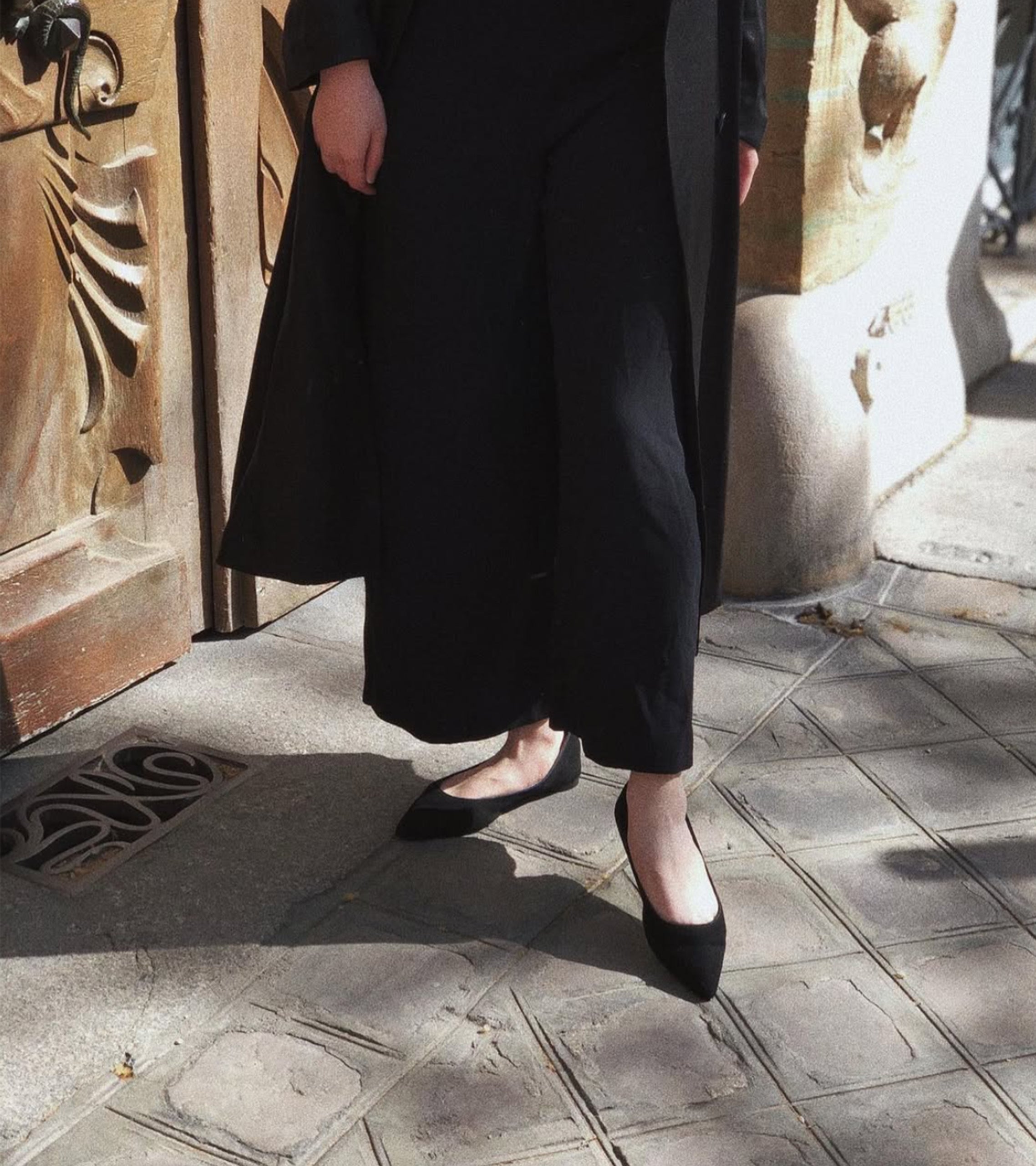 Person wearing a long black coat and black shoes on a stone pavement. #color_milano black