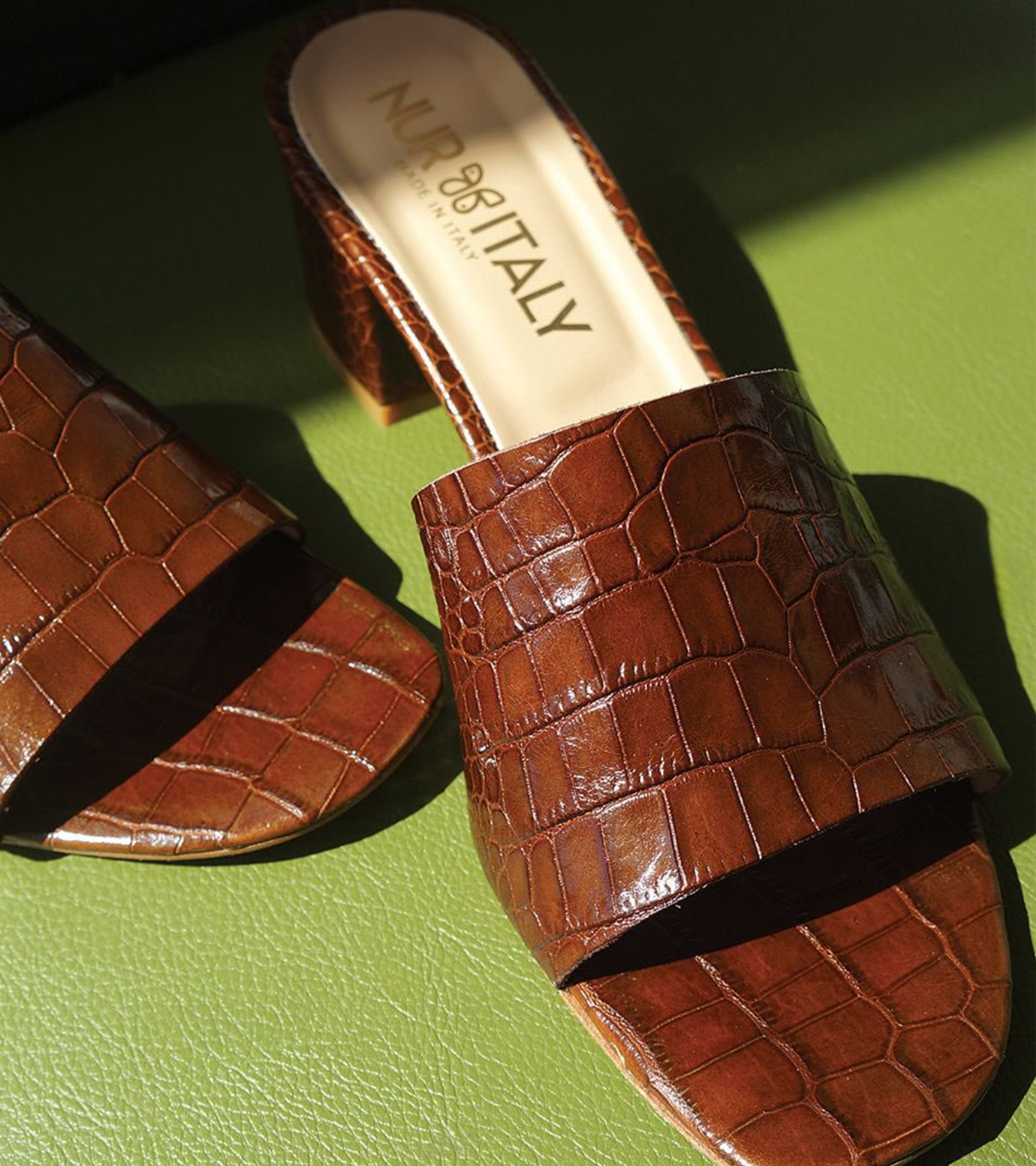 Brown crocodile-patterned slide sandals on a green surface with 'Nuria Italy' branding. #color_palermo tan